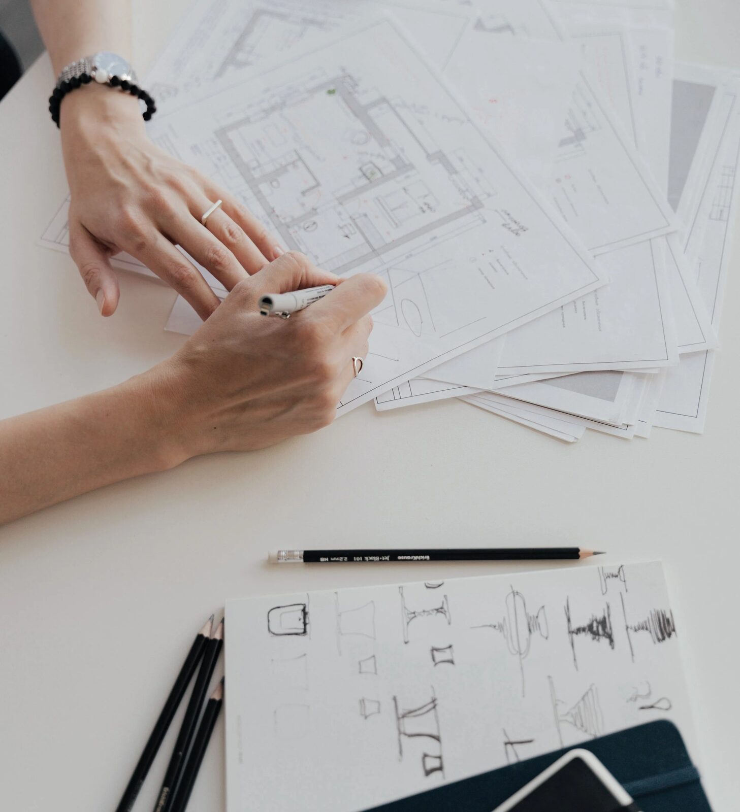 Mâini lucrând pe planuri de arhitectură, cu schițe de mobilier și creioane pe masă